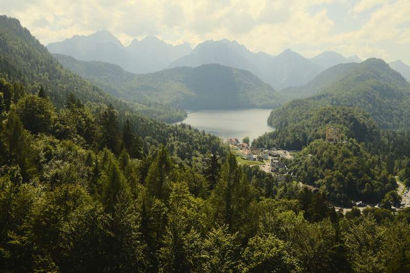 A summit view down to Alpsee, with a castle on the shore and the Alps fading out.