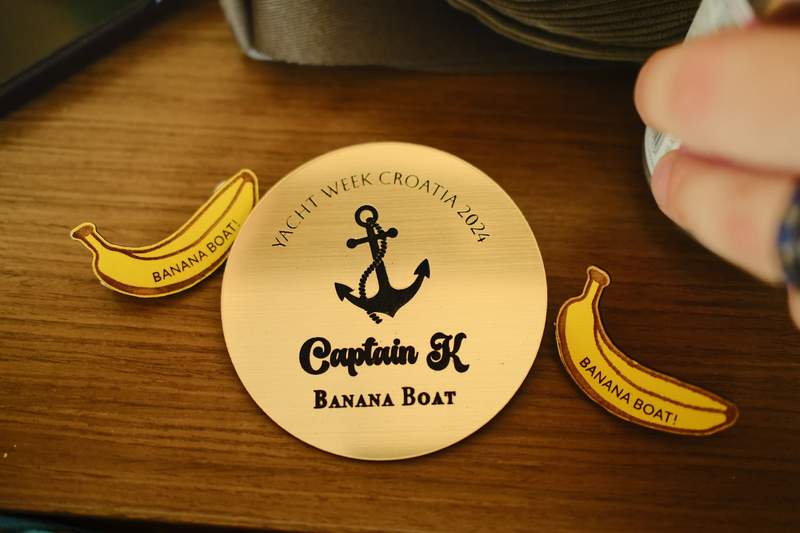 A hand holding a round Yacht Week skipper badge, with a boat deck and the sea behind it.
