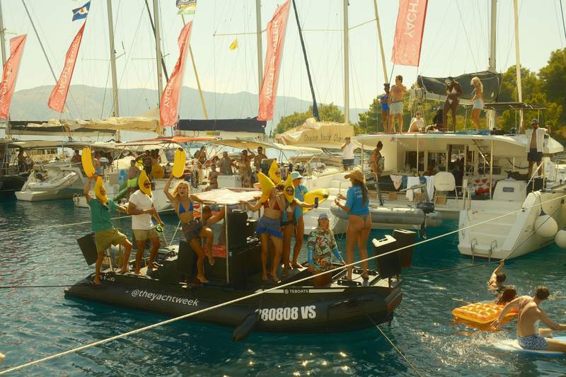 Catamarans and sailboats tied together around a boat with a DJ setup, with people swimming between the hulls.